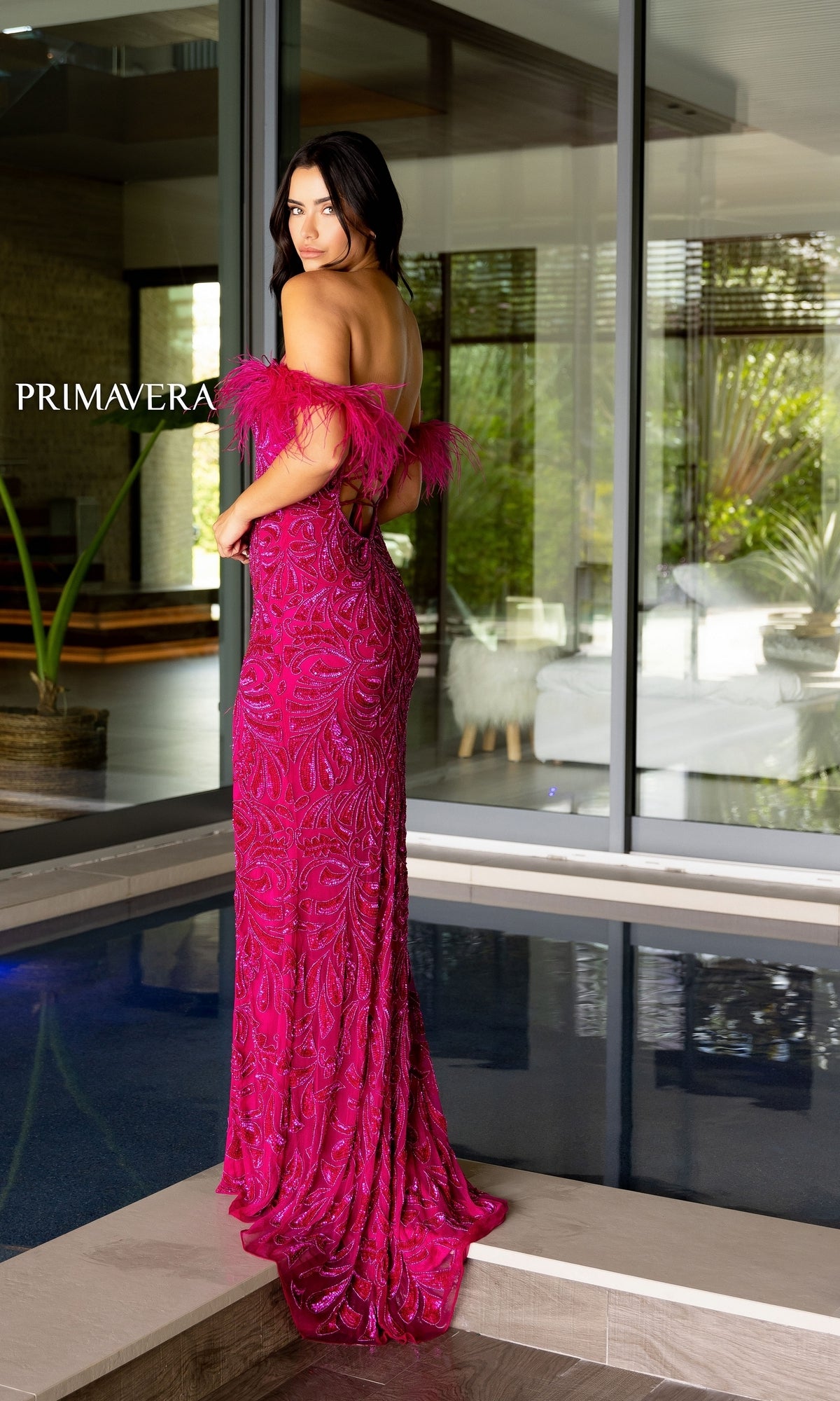 A woman stands indoors near a glass wall, facing away from the camera, wearing the pink Primavera Feather Off-Shoulder Long Prom Gown 4105 with feathered sleeves.